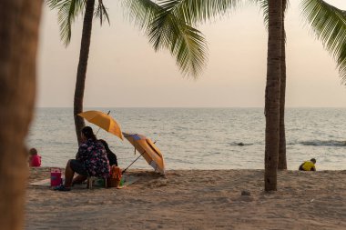 Chon Buri, Tayland - 26 Aralık 2018, kumlu sahilde uzanan birkaç uzun palmiye ağacının olduğu bir plaj sahnesi. İki kişi sarı bir şemsiyenin altında kumda oturuyor.