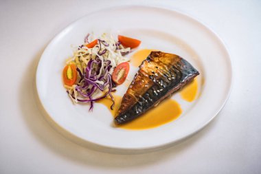 Beyaz tabakta teriyaki usulü ızgara uskumru ve açık turuncu-kahverengi teriyaki sosu var.