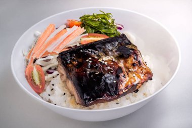 Beyaz, yuvarlak bir kasede, üzerinde teriyaki usulü ızgara balık olan bir porsiyon pirinç bulunur.