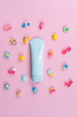 Pembe arka planda pastel mavi losyon tüpleri. Etrafı renkli plastik oyuncaklar, çiçekler ve diğer oyuncaklarla çevrili.