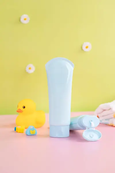Sarı plastik ördekli açık mavi bir losyon tüpü. Arka plan küçük beyaz çiçekli yumuşak bir yeşildir ve yüzey de bir pastel pembedir.