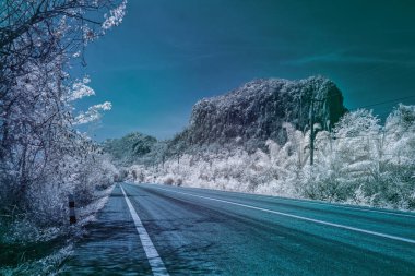Kızıl ötesi bir fotoğraf, berrak, canlı bir gökyüzünün altında yemyeşil bir bitki örtüsüyle kaplı büyük, kayalık bir tepeye uzanan sakin, boş bir asfalt yolu tasvir eder.
