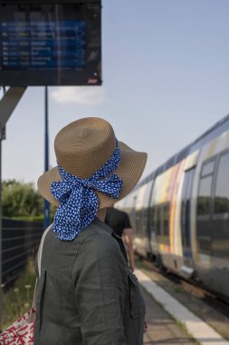 Goxwiller tren istasyonu. Mavi kurdeleli hasır şapkalı ayakta duran bir kadın istasyonda tren bekliyor.
