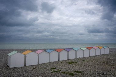 Le Treport, France - 09 16 2024:  View of white beach cabins with colorful roofs, wooden paths, pebble beach, blue sea and a cloudy sky clipart