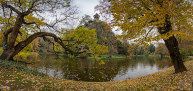 Paris, Fransa - 116 2024: Park Buttes Chaumont. Parkın orta kısmını yaya köprüsü, Belvedere adası, Sibyl Tapınağı, göl, fevkalade ağaçlar ve sonbahar renkleriyle bitki örtüsü ile görün.