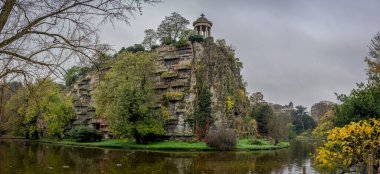 Paris, Fransa - 11 11 11 2024: Park Buttes Chaumont. Parkın orta kısmını yaya köprüsü, Belvedere adası, Sibyl Tapınağı, göl, fevkalade ağaçlar ve sonbahar renkleriyle bitki örtüsü ile görün.