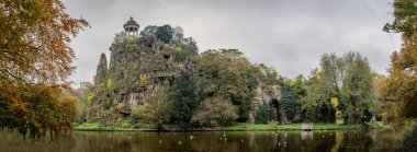Paris, Fransa - 11 11 11 2024: Park Buttes Chaumont. Parkın orta kısmını yaya köprüsü, Belvedere adası, Sibyl Tapınağı, göl, fevkalade ağaçlar ve sonbahar renkleriyle bitki örtüsü ile görün.