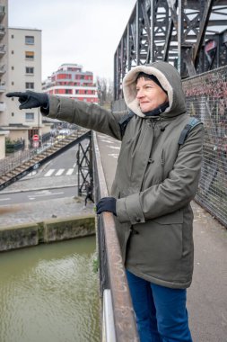Haki kapüşonlu ceket giyen olgun beyaz bir kadın köprüde durur ve kışın ufka bakar.