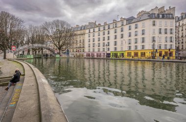 Paris, Fransa - 02 23 2022: Canal Saint-Martin. Beyaz ve kıvrımlı binaların panoramik manzarası, renkli depoların bir köprüsü ve suya yansıyan ağaçlar.