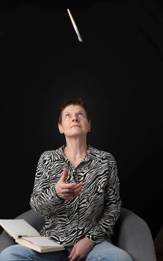 Stüdyo fotoğrafı. Gülümseyen olgun bir kadın bir çizim defterindeki koltukta oturuyor.