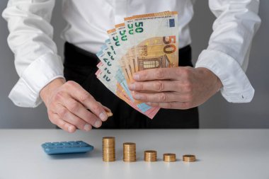 Yakından bakan adamın eli altın sikkeleri beşin arasına koyuyor, diğer eli de banknot bileziğini ve yanındaki hesap makinesini beyaz bir masanın üzerine koyuyor.