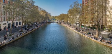 Paris, Fransa - 04 05 2025: Canal Saint-Martin. İnsanların panoramik manzarası, apartmanlar, suya yansıyan bir köprü ve ağaçlar.