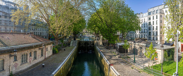 Paris, France - 04 05 2025: Canal Saint-Martin. Panoramic view of people sited, apartment buildings, a bridge and trees reflecting on the water
