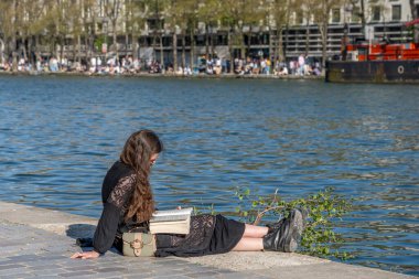 Paris, Fransa - 04 05 2025: La Villette Havzası. Güneş gözlüğü takan esmer beyaz bir kadın Qay 'de oturmuş kitap okuyor.