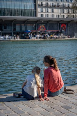 Paris, Fransa - 04 05 2025: La Villette Havzası. Bir bardak birası olan esmer beyaz bir kadın ve genç sarışın kızı Qay 'de oturuyorlar.