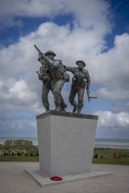 Vers-Sur-Mer, Fransa - 05 16 2025: British Normandy Memorial. Üç silahlı İngiliz askeri, arka planda altın plaj ve bayraklar olan bir çeliğin üzerinde duruyor ve koşuyor.
