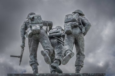Vers-Sur-Mer, Fransa - 05 16 2025: British Normandy Memorial. Üç silahlı İngiliz askeri, arka planda gri bulutlu gökyüzü ve bayraklarla bir stelin üzerinde duruyor ve koşuyor.