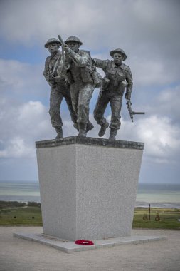 Vers-Sur-Mer, Fransa - 05 16 2025: British Normandy Memorial. Üç silahlı İngiliz askeri, arka planda altın plaj ve bayraklar olan bir çeliğin üzerinde duruyor ve koşuyor.