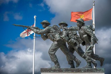 Vers-Sur-Mer, Fransa - 05 16 2025: British Normandy Memorial. Üç silahlı İngiliz askeri, arka planda altın plaj ve bayraklar olan bir çeliğin üzerinde duruyor ve koşuyor.