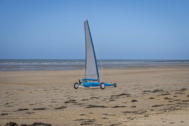 Asnelles-Sur-Mer, Fransa - 05: 16 2025: Kum ve deniz arasında sahil boyunca bir yelkenli tarafından itilen beyaz ve mavi bir kara yatı manzarası