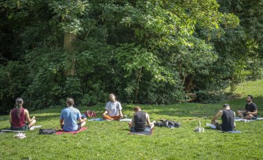Paris, Fransa - 06 14 2025: Buttes-Chaumont Parkı. Çimlerin üzerinde oturup yoga ve bitki örtüsü yapan bir grup insanın görüntüsü.