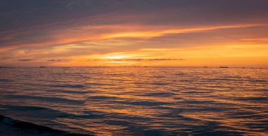 Denizin panoramik manzarası, dalgalar, yapay liman ve iskele boyunca gün batımında renkli bulutlu gökyüzü.