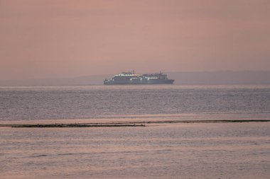 Luc-Sur-Mer, Fransa - 08 10 2025: Deniz üzerinde gün doğumuyla birlikte bir feribot görüntüsü ve gün doğumunda renkli ışığın yansımaları