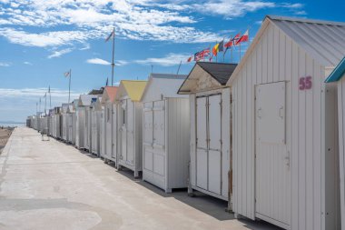 Langrune-Sur-Mer, Fransa - 08 07: 2025: Beyaz plaj kulübelerinin panoramik manzarası renkli çatılar, ulusal bayraklar ve deniz kıyısı boyunca mavi bulutlu bir gökyüzü