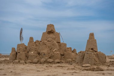 Kumsalda bir kumdan kale, kuş tüyü ve arkasında mavi bir bulut var.