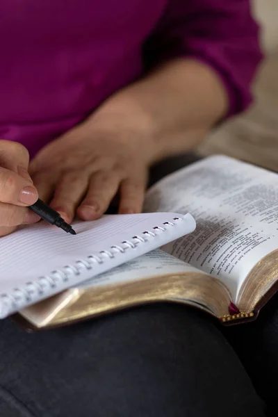 Senior Christian woman writing in a notebook with an open Holy Bible Book in her lap. Vertical shot, a close-up. Selective focus. Writing biblical verses, reading and studying Scriptures concept.