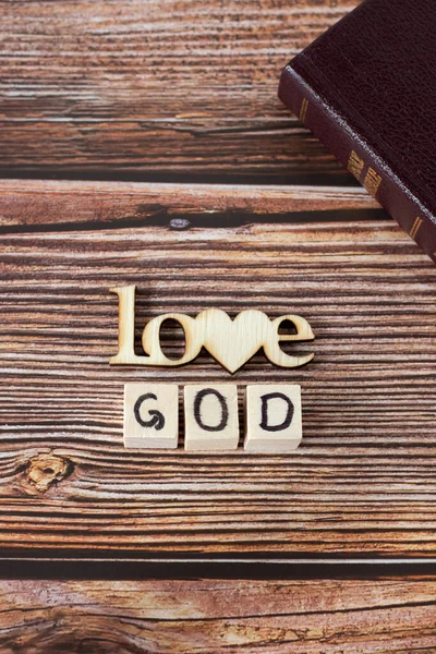 爱上帝的文字是用木制立方体写的 有心形的字母写在简朴的背景上 并附有合上的圣经 垂直射击 顶部视图 基督教圣经概念 — 图库照片