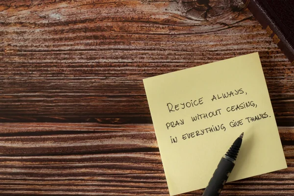 Stock image Rejoice always, pray without ceasing, give thanks to God Jesus Christ, handwritten new testament verse (Thessalonians 5:16) on note with pen and holy bible book on wooden table. Copy space, top view.