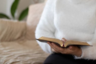 Christian woman holding open holy bible book and reading biblical verses at home. A closeup. Selective focus. Biblical concept of Studying the Scripture.