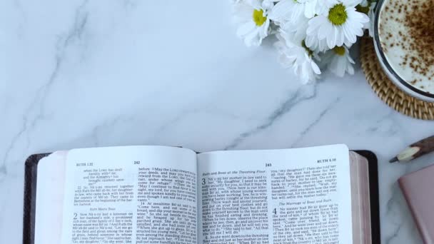 手翻开圣经书的翻页 上面有一杯咖啡和白色大理石背景的花朵 顶部视图 基督教教育 在家里学习圣经概念 — 图库视频影像