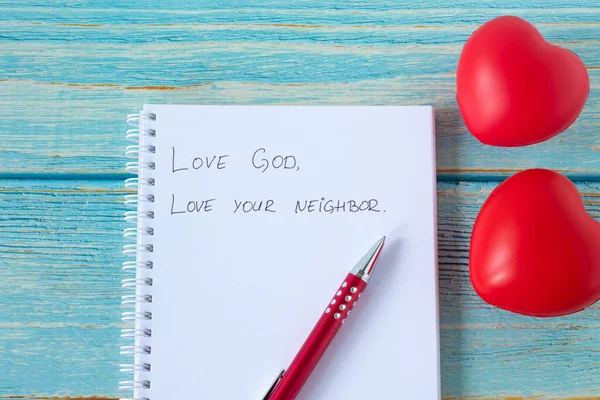 Love God and your neighbor handwritten quote in notebook with pen and red heart shapes on wooden background. Christian faith, obedience to Jesus Christ, bible\'s great commandment, biblical concept.