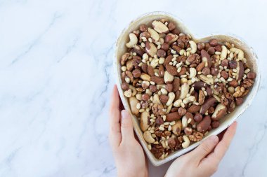 Karışık fındık (fındık, badem, ceviz, Brezilya ve çam fıstığı), beyaz arka planda kadın eli ile kalp şeklinde bir tabakta bulunur. Üst Manzara. Boşluğu kopyala.