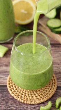 Ahşap masaya, kereviz, salatalık ve ıspanakla birlikte yeşil detoks smoothie döküyorum. Kahvaltıda sağlıklı organik karışım. Dikey video, yakın plan.
