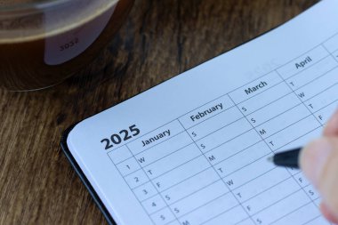 Ocak 2025 takvimi, ahşap masa üzerinde kahve fincanı olan bir not defteri. Aylık tarih düzenleyici, program şablonu. Yakın plan..