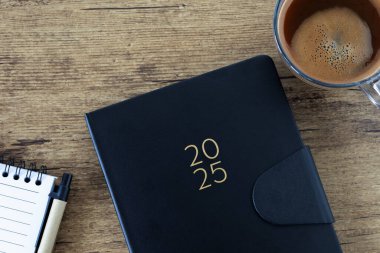 2025 yılbaşı not defterinde bir fincan kahve ve ahşap arka plan. Üst masa manzaralı. Takvim planlayıcısı, organizatör, iş etkinliği takvimi kavramı.