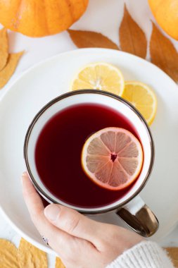 Kadın eli, sonbahar yaprakları ve balkabağıyla beyaz mermer masada limonlu bir fincan amber çayı tutuyor. Üst Manzara. Kadın ev yapımı sıcak içecekle rahatlıyor..