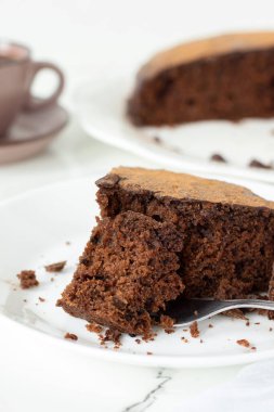Arkaplanda kahve fincanı olan beyaz bir tabakta çikolatalı kek dilimi. Yakın plan. Seçici odaklanma. Ev yapımı vejetaryen tatlı, kakao ve keçiboynuzu tozu..