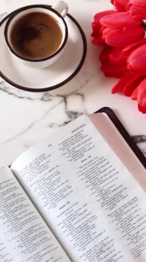 El, kutsal İncil 'in sayfalarını bir fincan kahve ve beyaz mermer masada kırmızı lalelerle çeviriyor. Dikey video. Üst Manzara. Hristiyan yazıtları okumak, İncil kavramı.