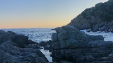 Güneş batarken üzerine deniz dalgaları düşen bir uçurumun panoramik görüntüsü.