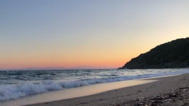 Sahilde gün batımının videosu. Sahilde dalgalar ve arka planda dağlar.