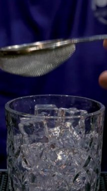 Bir barmenin alkollü kokteyli filtrelediği buzlu dikey yakın çekim bardağı. Gece hayatı, alkollü içkiler, kulüpler, barmenlik mesleği.