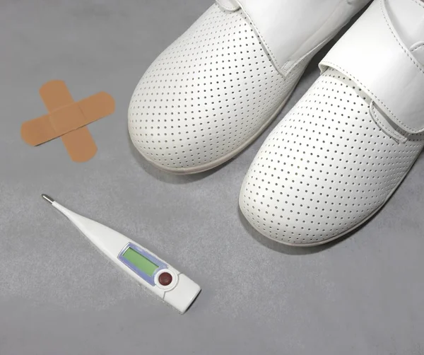 Medical shoes, medical concept, pair of white professional ventilated work clogs isolated on gray and band aid and thermometer