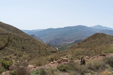 Endülüs 'ün güneyindeki dağ manzarası, arazi sarp, çalılar ve ağaçlar var, dağ yamacından geçen bir yol, gökyüzü açık.