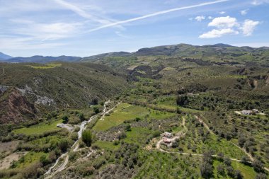 Granada 'nın güneyindeki dağlık arazi, bir vadi ve bir nehir var, birkaç çiftlik evi var, birkaç zeytin ve badem ağacı var, gökyüzünde bulutlar var.