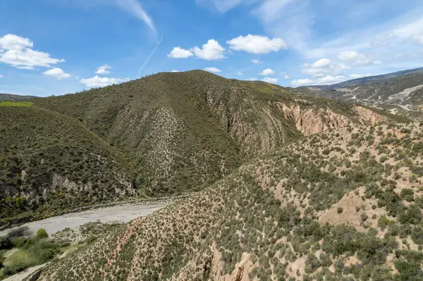 Granada 'nın güneyindeki dağlık arazi, dağlarda çalılar, dağların arasından geçen kuru bir nehir, gökyüzünde bulutlar var.