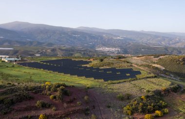Granada 'nın güneyinde Photovoltaic panel tesisi, hemen yanında bir çiftlik var, dağlarda birkaç köy var, ağaçlar ve çalılar var, gökyüzü açık.